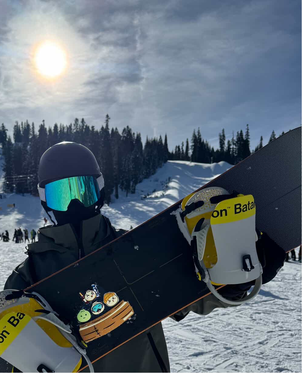 Person holding a snowboard with a mountain and skiers in the background